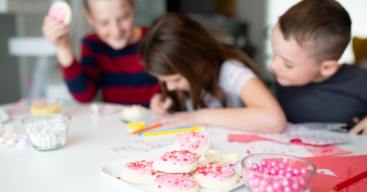 "Three children decorating Valentine's Day cookies at a table filled with frosting, sprinkles, and candies. The kids are laughing and focused on their creations, capturing a fun and festive moment