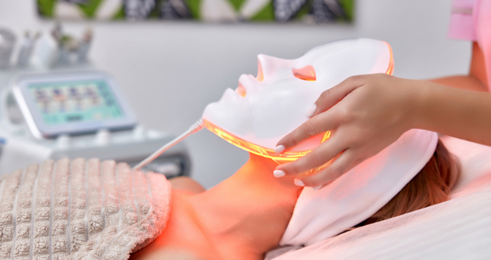 A person lying down during a skincare treatment with a white LED mask emitting red light. The setting is a modern spa with soft lighting and a calm atmosphere, featuring skincare equipment in the background
