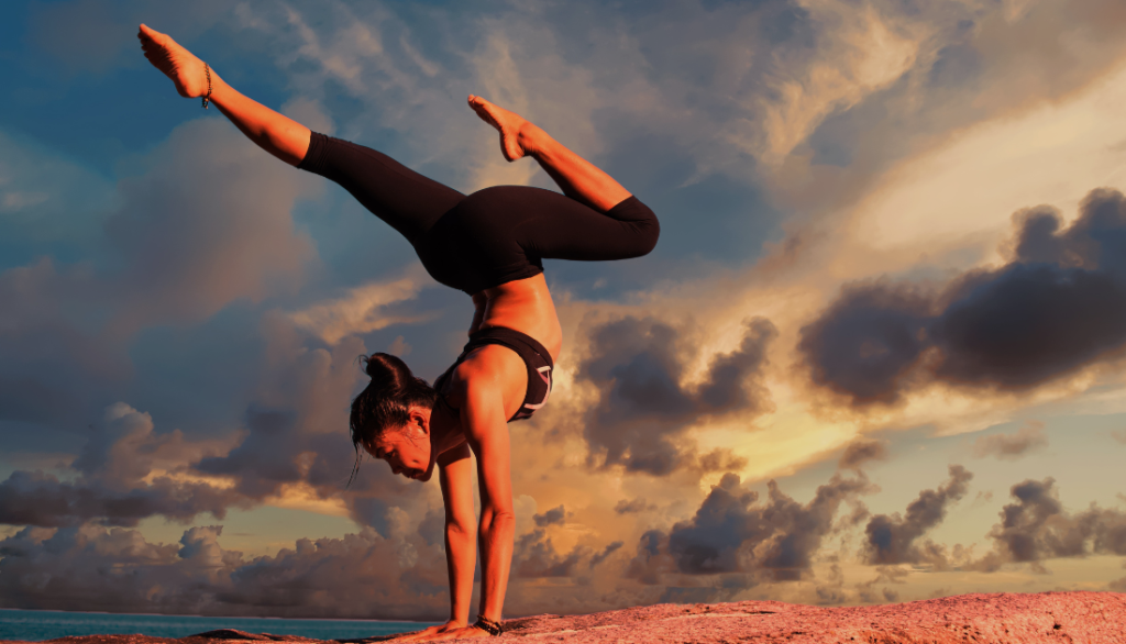 A woman performing a handstand on a rocky surface during a dramatic sunset. She is dressed in black athletic wear, balancing gracefully against a backdrop of vibrant orange and blue skies with scattered clouds. The scene conveys strength, focus, and serenity.