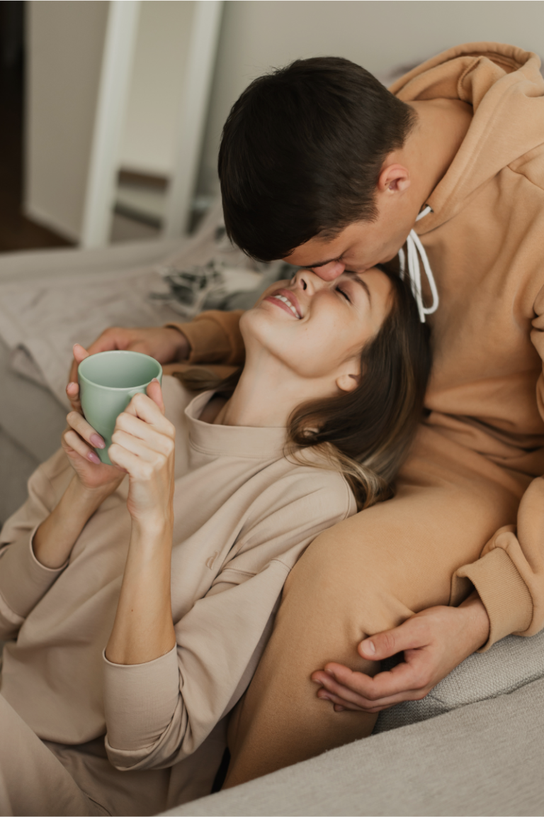 Couple sharing a tender moment, with a woman holding a green coffee mug while smiling warmly as her partner kisses her forehead. Both are dressed in cozy, neutral-toned loungewear, enjoying a relaxed and intimate setting at home.