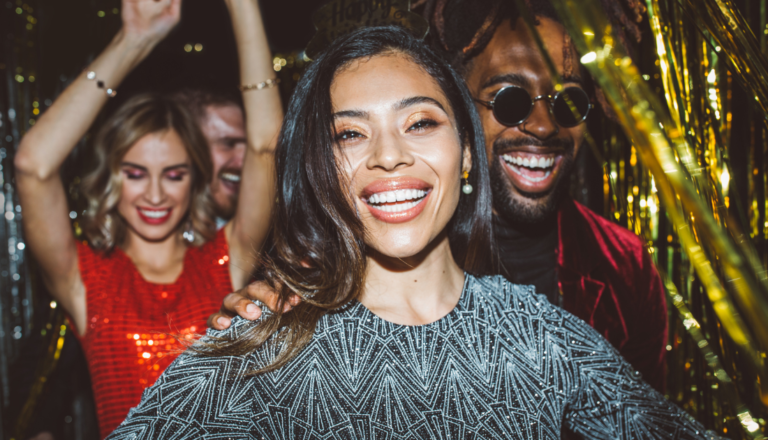 A group of friends celebrating at a festive party. A smiling woman in a sparkly silver dress is in focus, surrounded by others laughing and enjoying themselves. Gold tinsel decorates the background.
