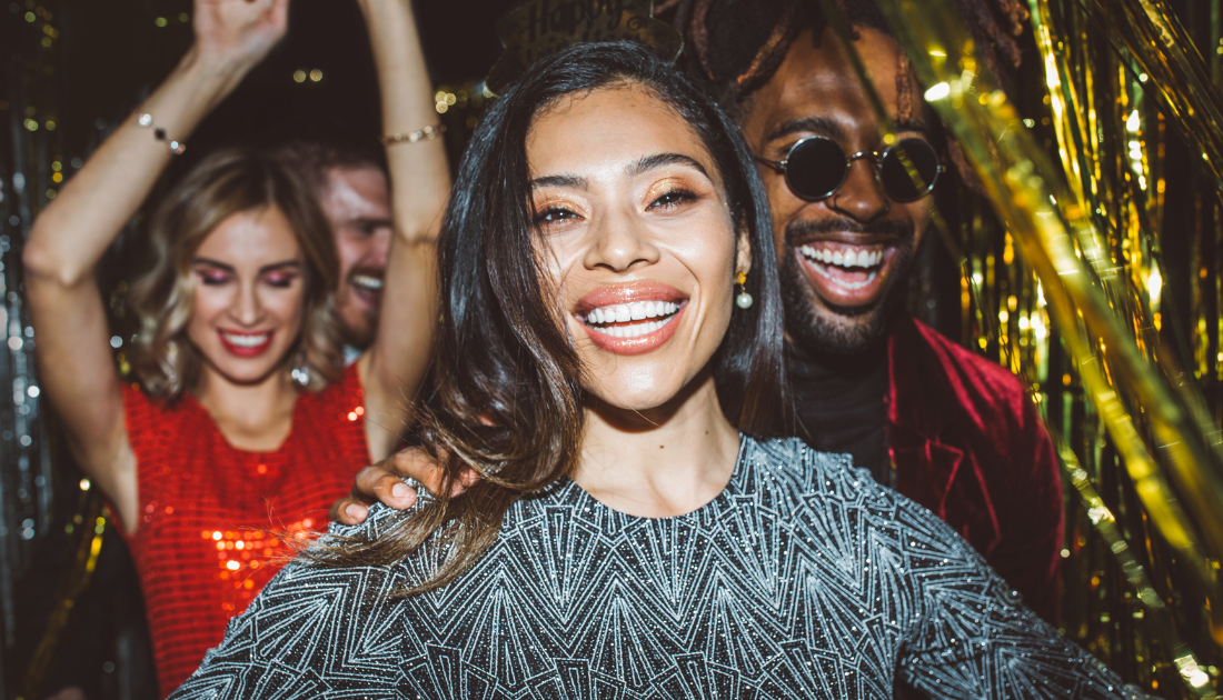 A group of friends celebrating at a festive party. A smiling woman in a sparkly silver dress is in focus, surrounded by others laughing and enjoying themselves. Gold tinsel decorates the background.