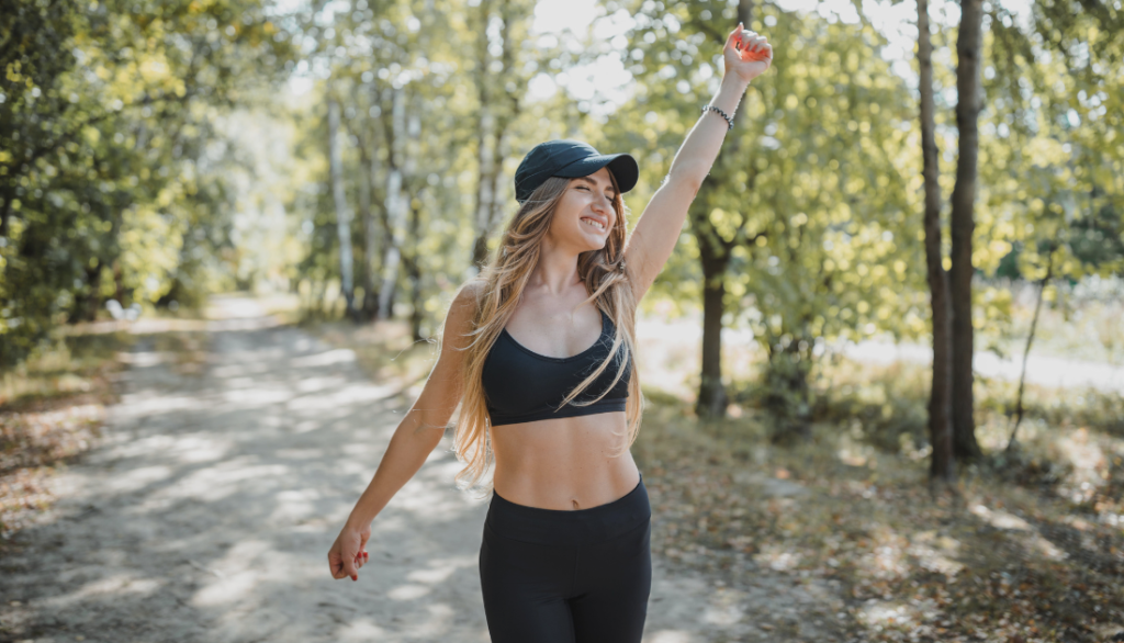  A cheerful woman in a black sports bra and cap raising her arm triumphantly during a walk in a lush forest. She is smiling and appears energized, surrounded by tall trees and a sunlit path