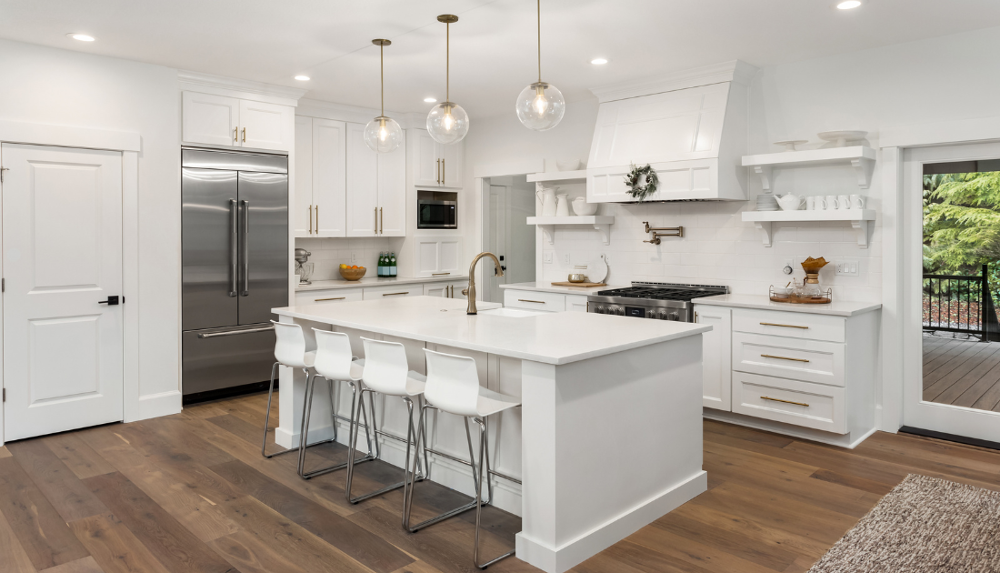 A bright, spacious modern kitchen with white cabinetry and a large center island. The kitchen features gold hardware, pendant lights, and an open shelf design. A stainless steel refrigerator and wide-plank wooden flooring complete the look.