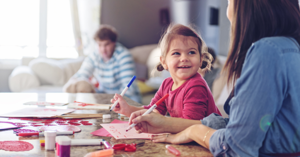 A cheerful young girl crafting Valentine's Day cards at a table with colorful markers and supplies, smiling at her mom as they create together. A family member relaxes in the background, creating a cozy and heartwarming scene