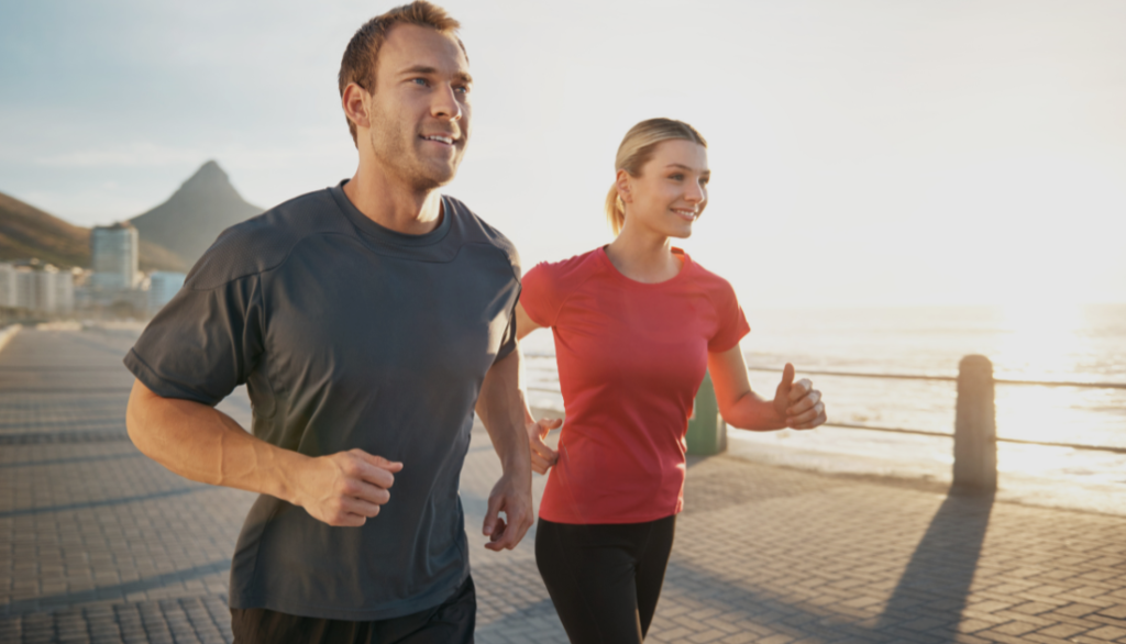 A man and woman jogging along a scenic waterfront promenade at sunrise. They are smiling, dressed in workout clothes, and surrounded by a vibrant landscape featuring a mountain and the ocean.
