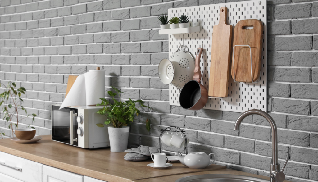 A modern kitchen wall with gray brick design featuring a pegboard organizer. Various kitchen tools, cutting boards, and a small plant are neatly arranged. A microwave and paper towel holder sit on the wooden countertop.