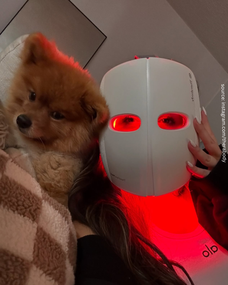 Person wearing a white LED light therapy mask emitting red light, holding a fluffy brown Pomeranian dog wrapped in a checkered blanket. The cozy setting emphasizes relaxation and self-care, with a modern and intimate ambiance.