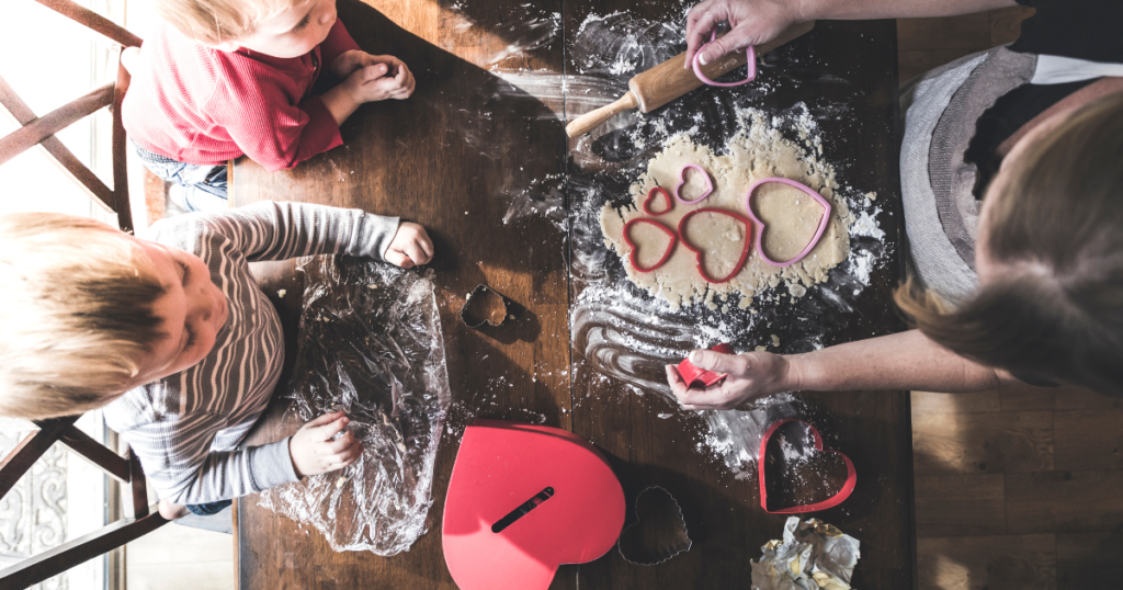 Top-down view of a family baking Valentine's Day cookies together, with heart-shaped cookie cutters and dough on a floured wooden table. Two young children eagerly watch and participate, creating a warm and festive moment