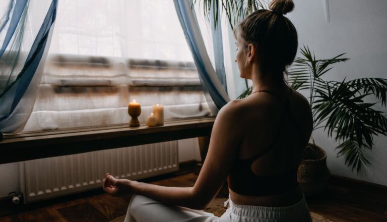 A woman sitting cross-legged in a peaceful meditation space, surrounded by soft natural light, candles, and indoor plants. She is dressed in comfortable activewear and facing a window with sheer curtains, creating a serene and calming atmosphere